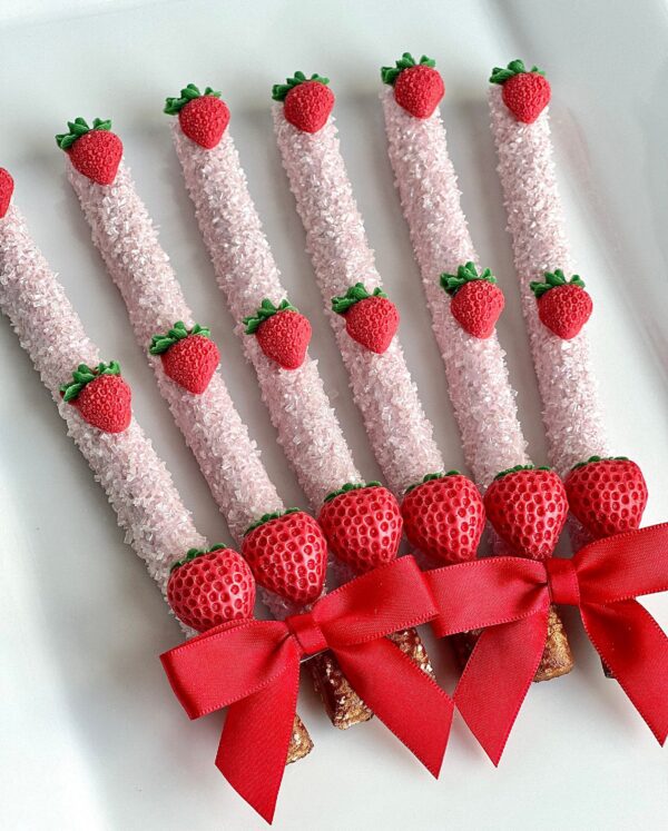 Six pretzel rods from the "12-Brilliant Strawberry Chocolate Covered Pretzel Rods" collection, elegantly coated in strawberry-themed toppings with small strawberry decorations and red bows, are arranged on a white plate.