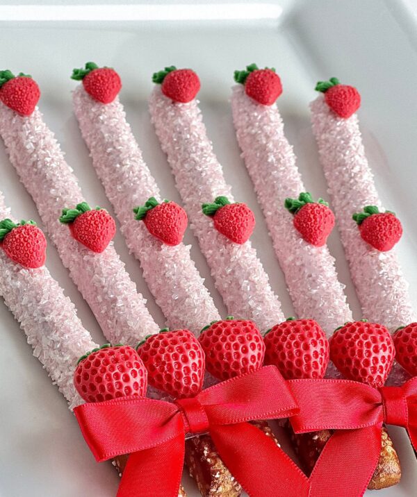 Rows of 12-Brilliant Strawberry Chocolate Covered Pretzel Rods are topped with small strawberry decorations on a white surface, perfect for birthdays, anniversaries, special occasions, or baby showers.