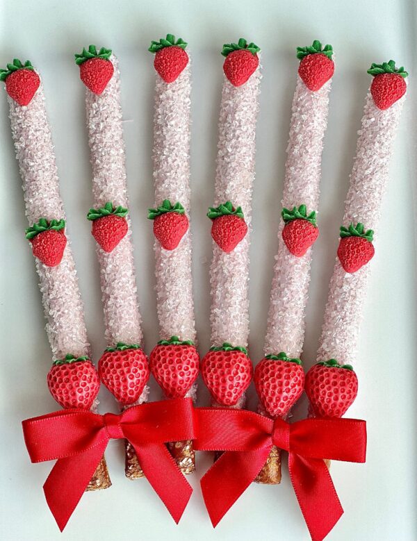 A set of six pretzel sticks adorned with pink icing, strawberry decorations, and red bows at the bottom.