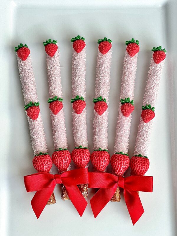 Six of the 12-Brilliant Strawberry Chocolate Covered Pretzel Rods are arranged vertically on a plate, featuring white chocolate coating, red strawberry decorations, and red bows at the bottom.