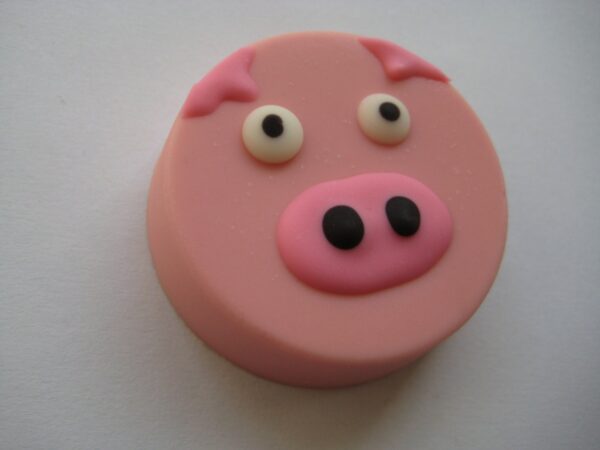 A pink pig face made of fondant with eyes, a nose, and ears rests on a white background as part of the 24-Pink Pig Chocolate Covered Oreo Favors.