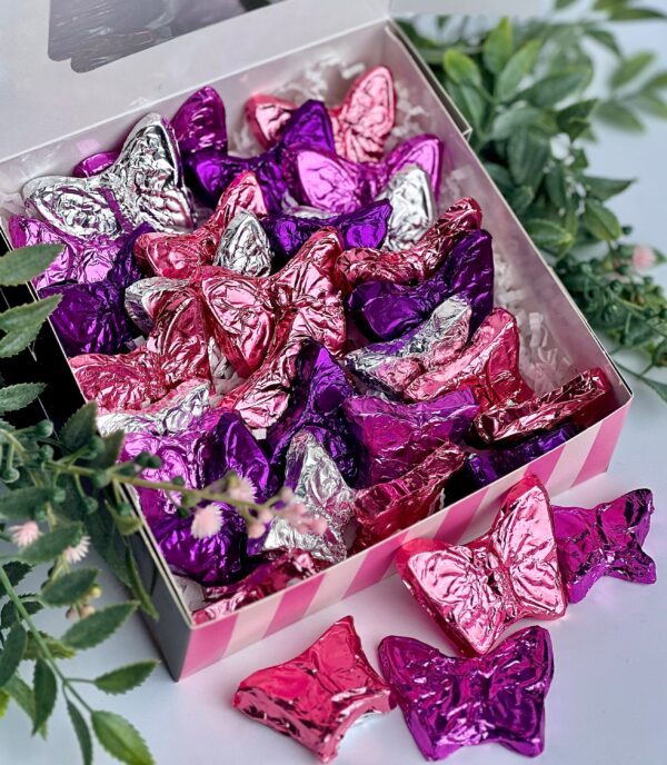 The Foil Wrapped Milk Chocolate Butterflies Gift Box, ideal for birthdays, anniversaries, or any occasion, features assorted purple, pink, and silver butterfly-shaped chocolates placed among green foliage on a white surface.