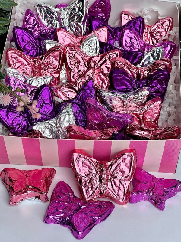 A gift box of milk chocolates wrapped in shiny pink, purple, and silver foil shaped like butterflies, with some displayed in front on a striped surface. Ideal for birthdays, anniversaries or any occasion.