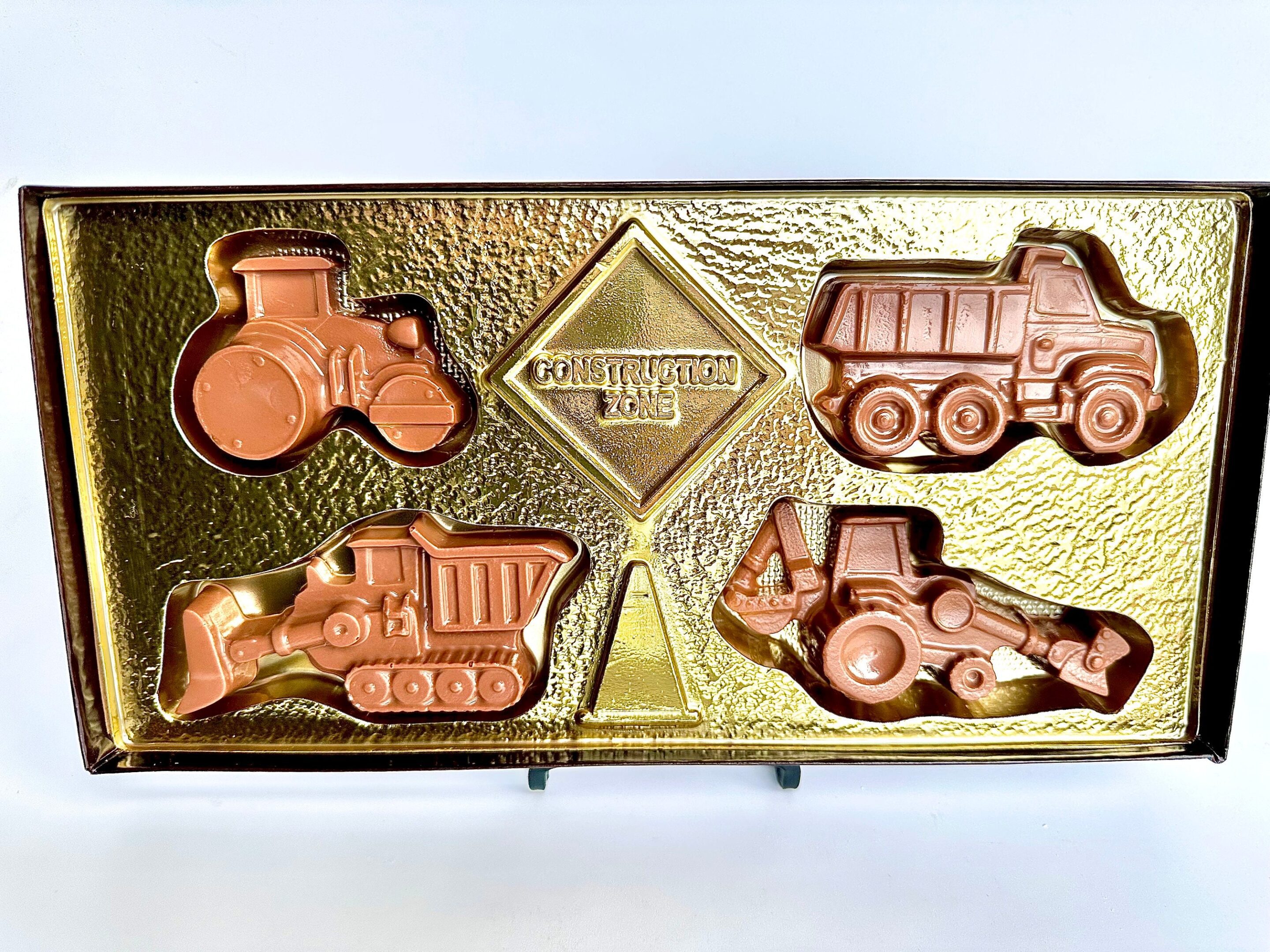 Box of chocolates shaped like construction vehicles, including a tractor, dump truck, front loader, and excavator, on a gold textured tray.