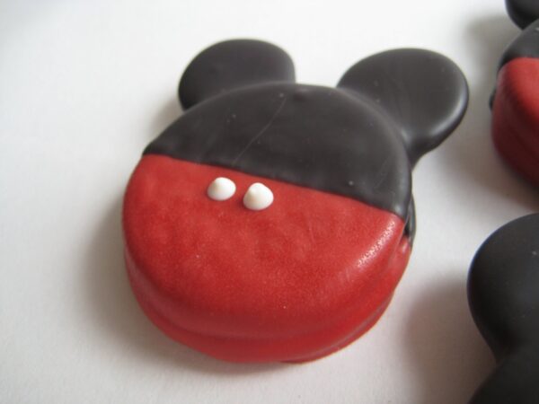 A cookie from the "12-Mickey Mouse Chocolate Covered Oreos" set, decorated with red and black icing to resemble mouse ears and featuring two white dots on a white surface.