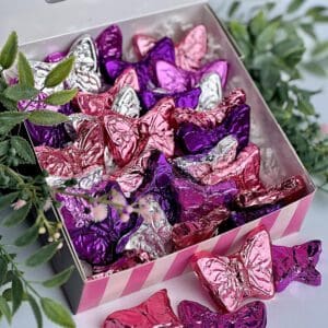 A gift box of milk chocolate butterflies wrapped in pink, purple, and silver foil with green leaves nearby.