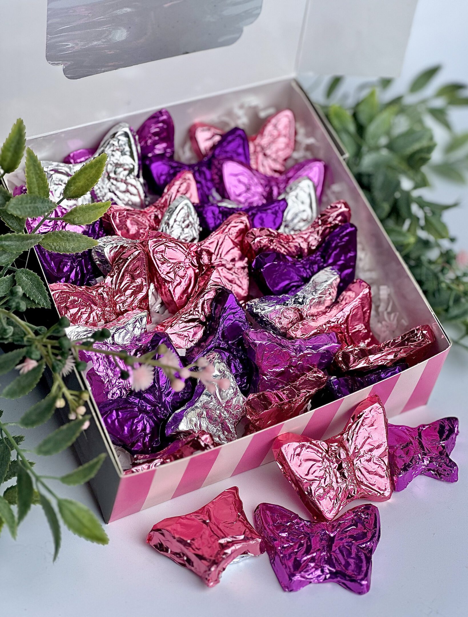 A gift box of milk chocolate butterflies wrapped in pink, purple, and silver foil with green leaves nearby.