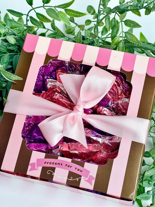 A gift box of pink and purple foil-wrapped milk chocolate butterflies, adorned with a pink ribbon, set against green leaves.