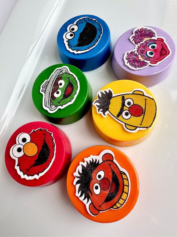 Colorful round chocolate-covered Oreo faces featuring cartoon designs in various colors like red, green, purple, yellow, orange, and blue are arranged on a white surface. Perfect as Sesame-themed party favors for birthdays or monster themes.