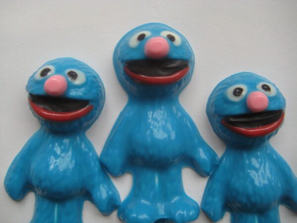 Three Grover-shaped chocolate lollipops feature blue cartoon characters with pink noses, wide eyes, and open mouths against a white background.