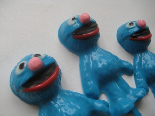 Three identical blue figures with large pink noses and wide smiles, resembling Grover chocolate lollipops, are lying on a surface.