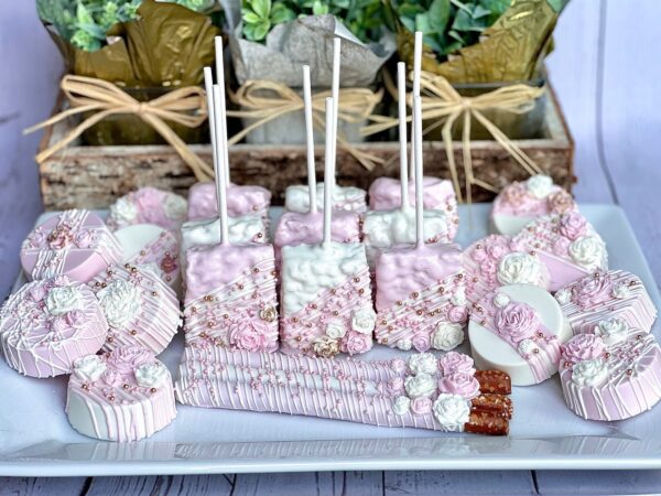 The Pink and White Chocolate Treat Party Bundle for wedding, birthday, or baby shower favors features artfully arranged pink and white desserts with floral and pearl embellishments, including cake pops, cookies, and chocolate-covered pretzels.