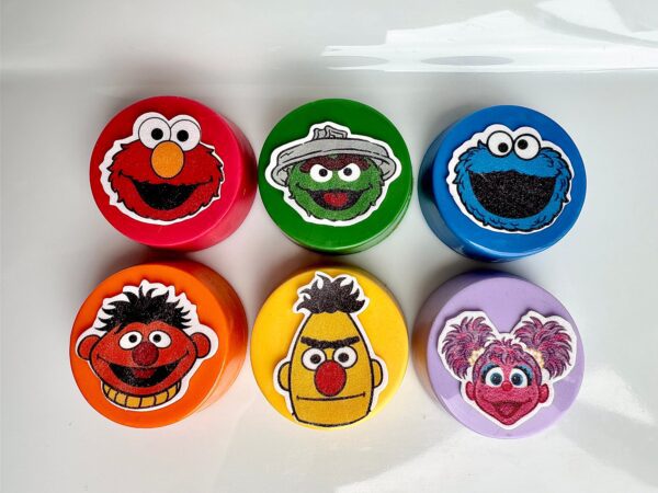 Six round containers with cartoon faces: red, green, blue, orange with a brown-haired character, yellow, and purple with a pink-haired character (Product: 12-Sesame Chocolate Covered Oreo Faces For Birthday Party/Monster Theme/Party Favors).