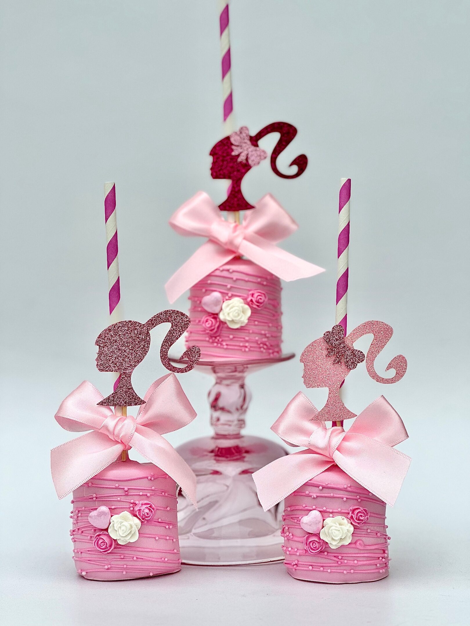 Three pink cake pops with silhouette toppers on striped sticks and ribbon bows are displayed on a glass stand.