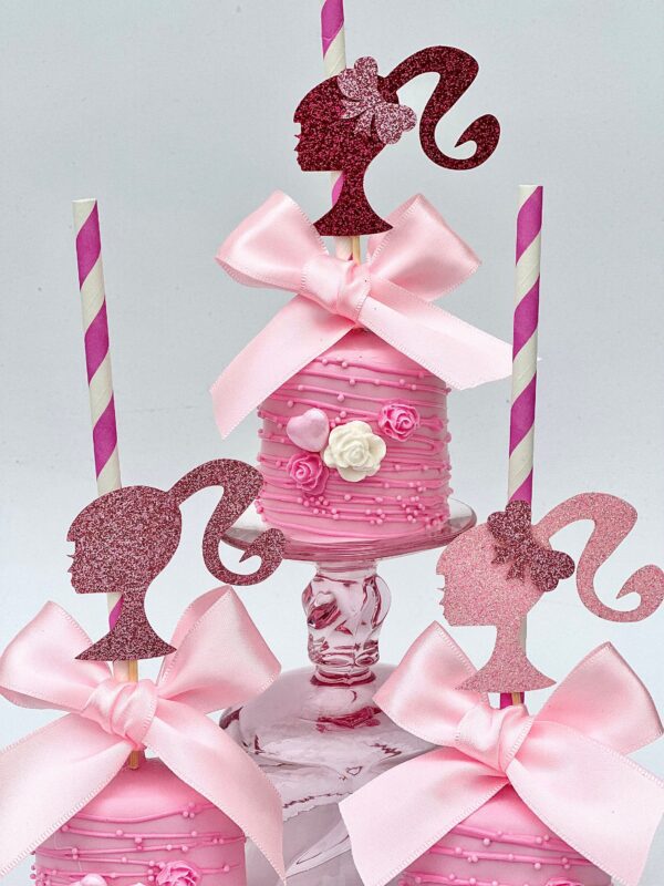 Three pink Barbi-themed chocolate-covered jumbo marshmallows decorated with silhouette toppers, pink bows, and striped straws are displayed on a stand.