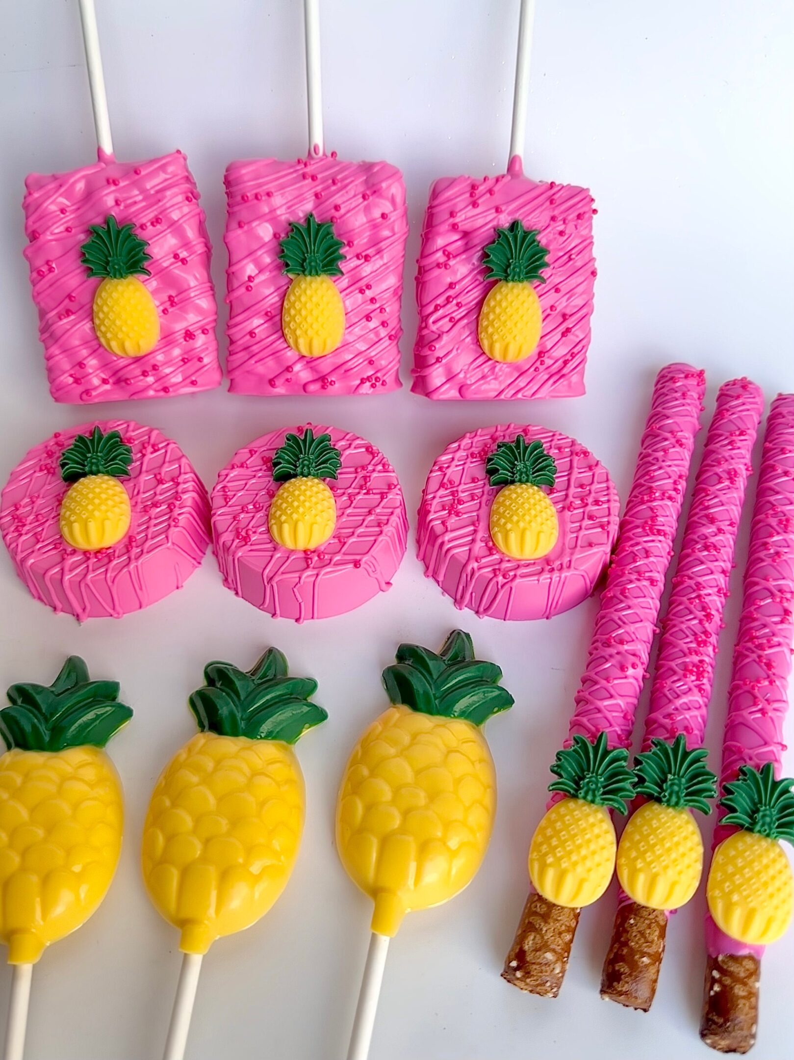 Assorted pink and yellow pineapple-themed treats: lollipops, chocolate-covered cookies, and pretzel sticks, all decorated with a pineapple motif.