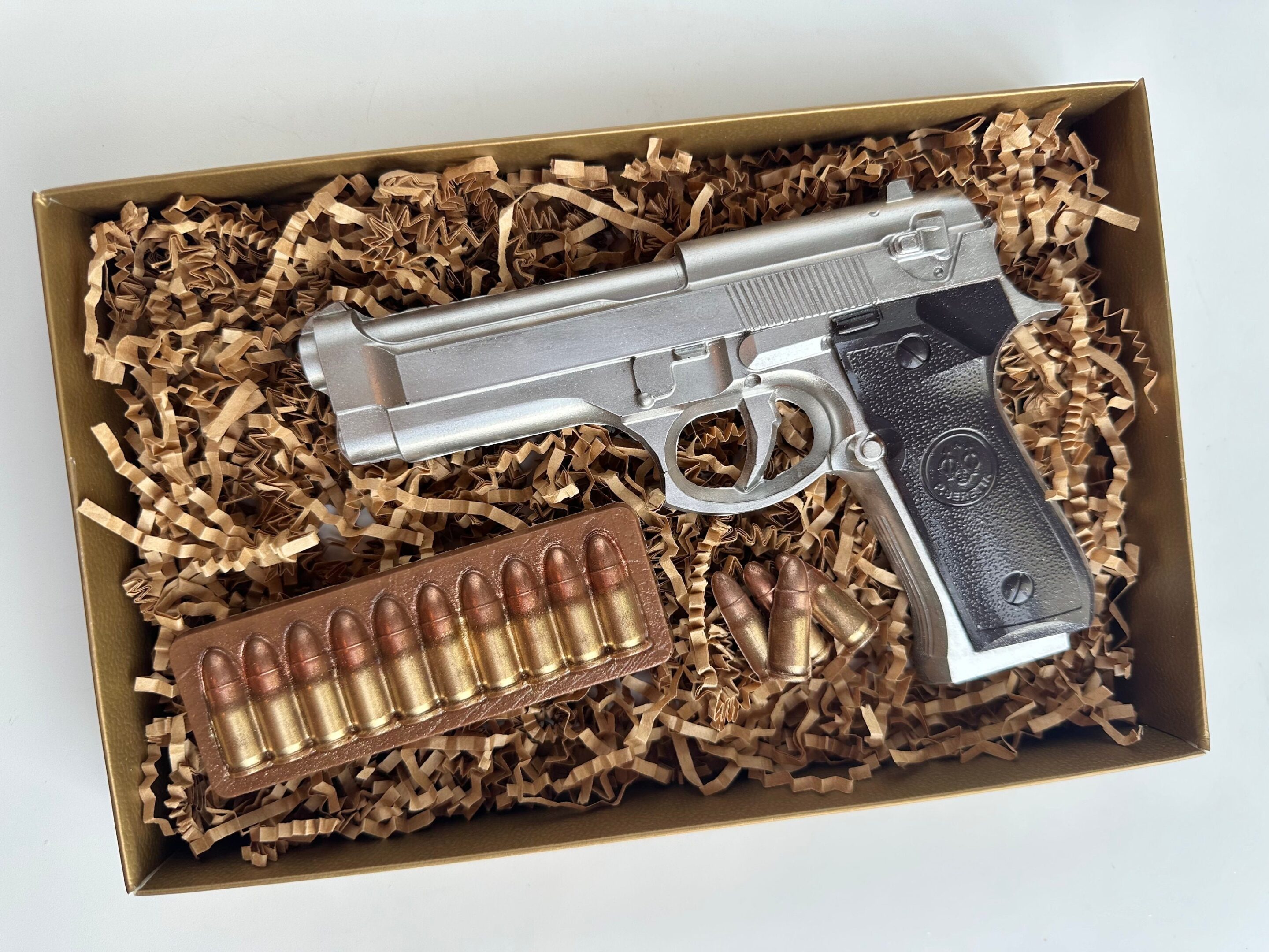 The Chocolate Hand Piece Boxed Gift includes a silver chocolate handgun with a black grip, nestled in brown crinkle paper, and paired with a tray of bullets and three loose rounds. Perfect for birthdays or as a gift for dads, sons, or police officers.