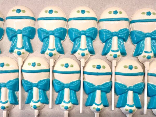 Baby blue and white cake pops shaped like rattles with bow designs, arranged in rows on a flat surface. Product: "12-Sweet Baby Boy White Chocolate Baby Rattle Lollipop Favors For Boy/Baby Shower Favors.