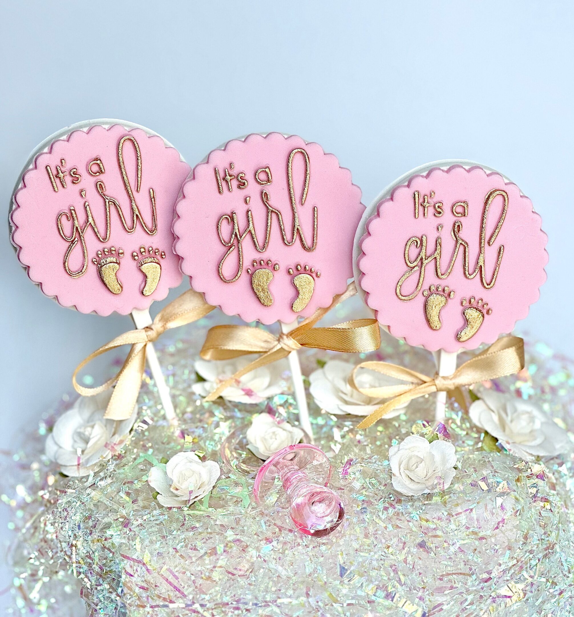 Three white chocolate lollipops from the "It’s A Girl" collection, featuring gold decorative elements, adorn a surface decorated with small white flowers and iridescent accents.