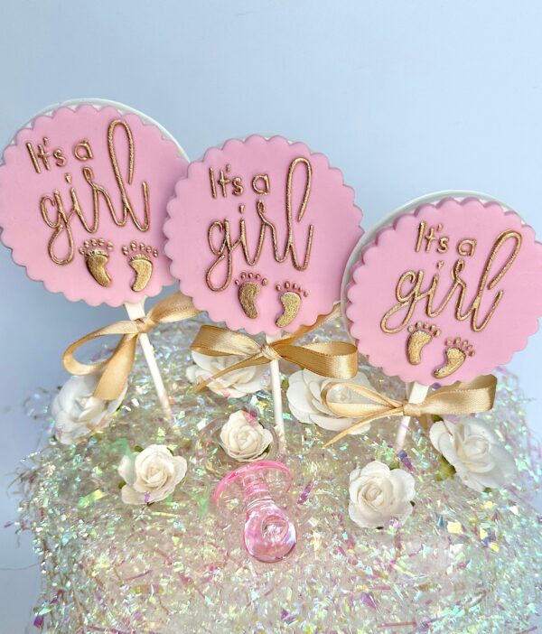 Three pink circular cake toppers with gold "It's a girl" text and baby footprints, adorned with gold bows, sit on a sparkly surface alongside white flowers and a small pink baby bottle. Product: 12-It’s A Girl White Chocolate Lollipops for Baby Shower/Party Favors.