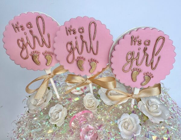 Three "It's a Girl" white chocolate lollipops with pink circular decorations, gold baby footprints, and ribbons on sticks, surrounded by roses on a sparkly surface.