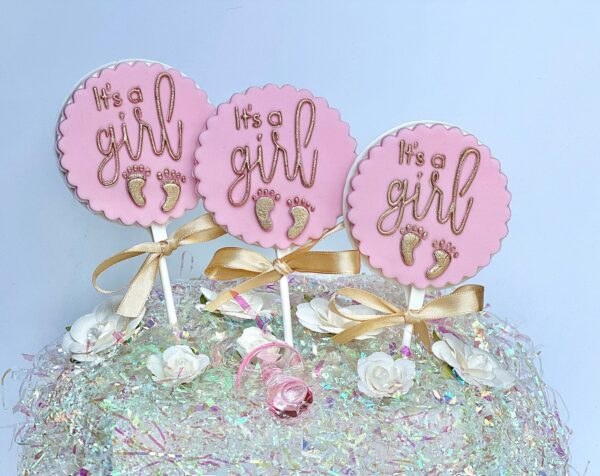 Three pink "It's a girl" cake toppers with gold text and footprints, garnished with gold bows, are set on a dazzlingly ornate cake featuring white roses alongside 12 "It's A Girl" white chocolate lollipops as party favors.