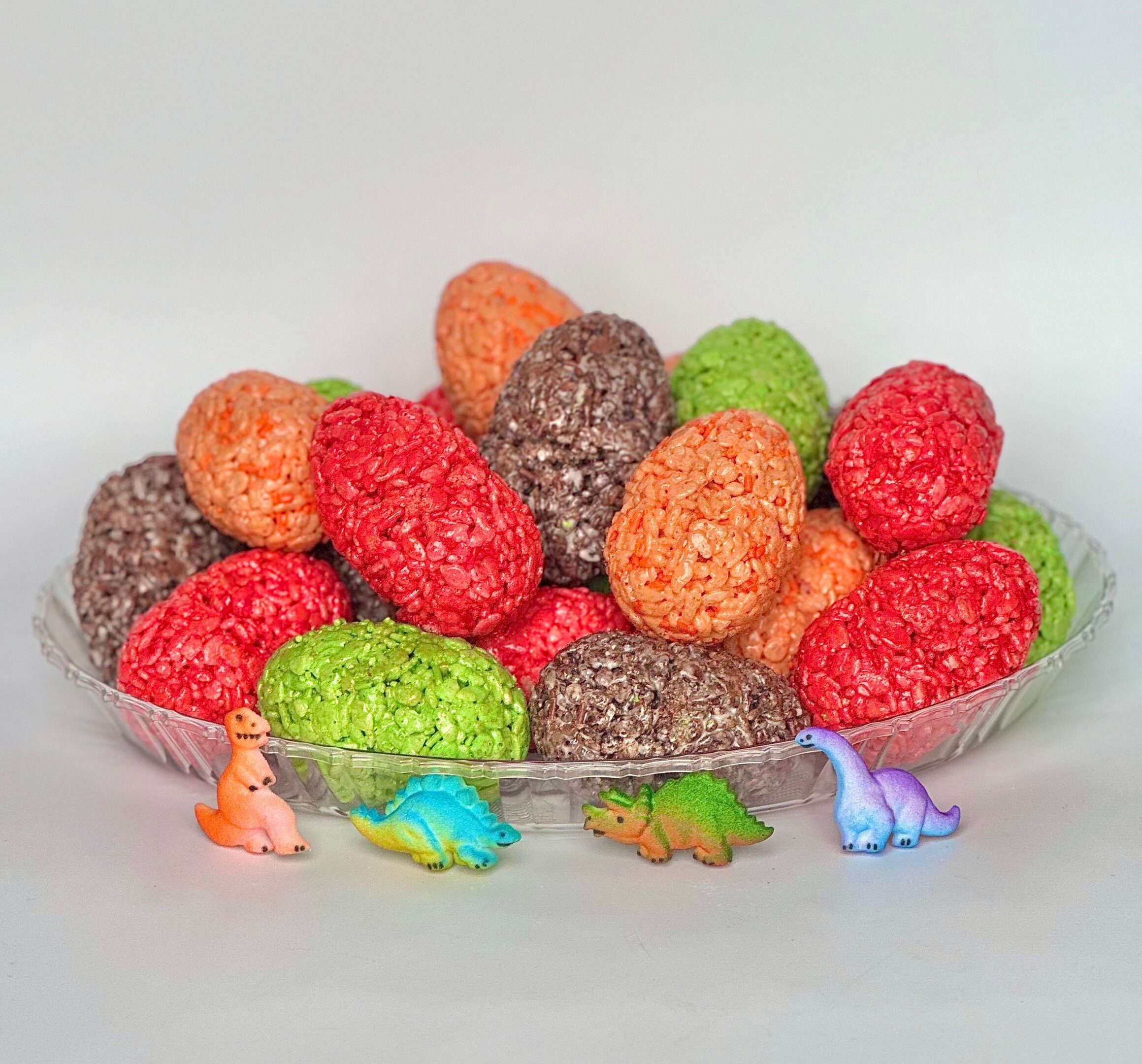 A glass bowl filled with 12 colorful, textured Dinosaur Egg Rice Krispie Treats for birthday or dinosaur-themed parties, with small dinosaur figurines placed in front.