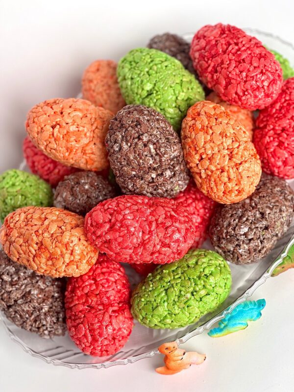 A glass dish displays colorful, oval-shaped "12-Dinosaur Egg Rice Krispie Treats" in red, green, brown, and orange with toy dinosaurs beside it.
