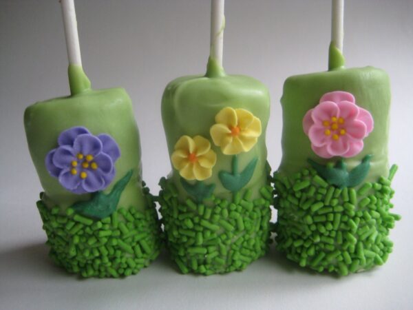 Three double marshmallow pops, from the "12-Flower Garden Themed Chocolate Covered Double Marshmallow Pops," are coated in green frosting, adorned with colorful flower designs, and topped with green base sprinkles.