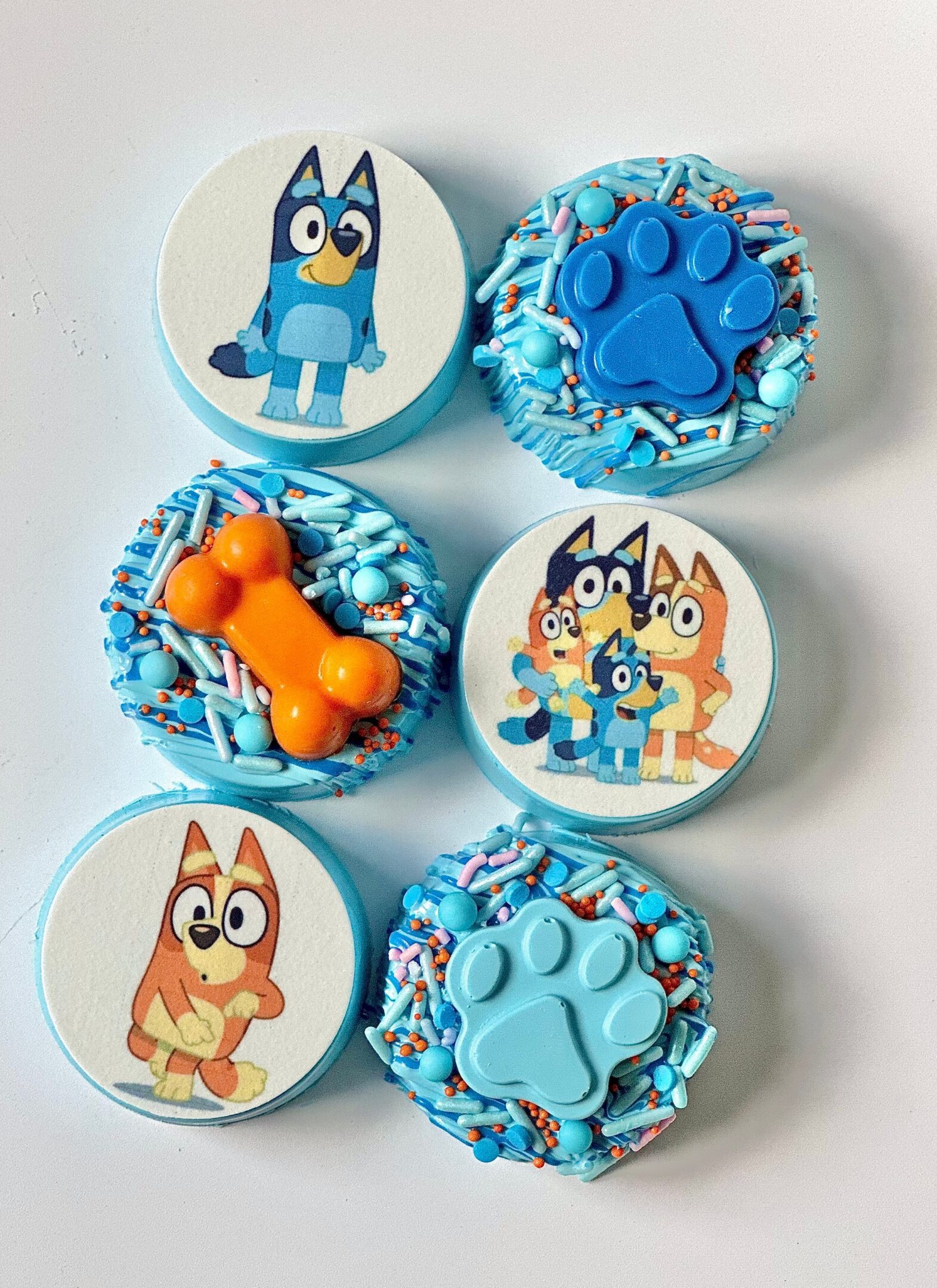 Indulge in 12 chocolate-covered Oreos designed with a "Bluey" theme, showcasing character images, blue paw prints, and orange bone shapes, all enhanced with blue and orange sprinkles. Perfect for birthday party favors!.