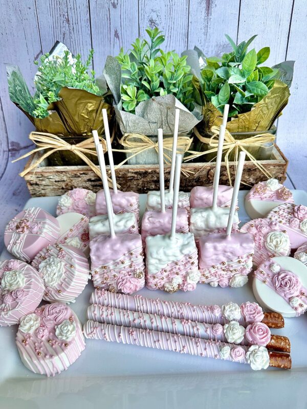 A display of the Pink and White Chocolate Treat Party Bundle, featuring decorated cake pops, pretzel sticks, and cookies, with small potted plants in the background.