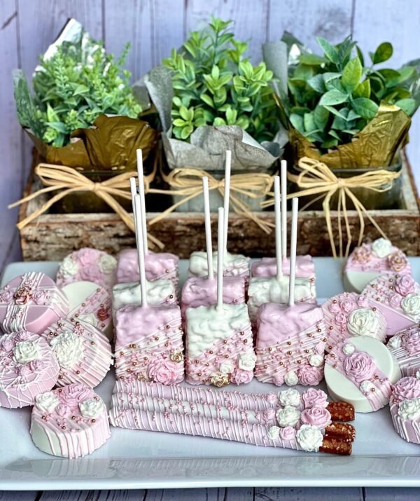 A Pink and White Chocolate Treat Party Bundle featuring decorated sweets like cake pops and pretzel rods is arranged on a white plate with green potted plants in the background, perfect for weddings, birthdays, or baby showers.