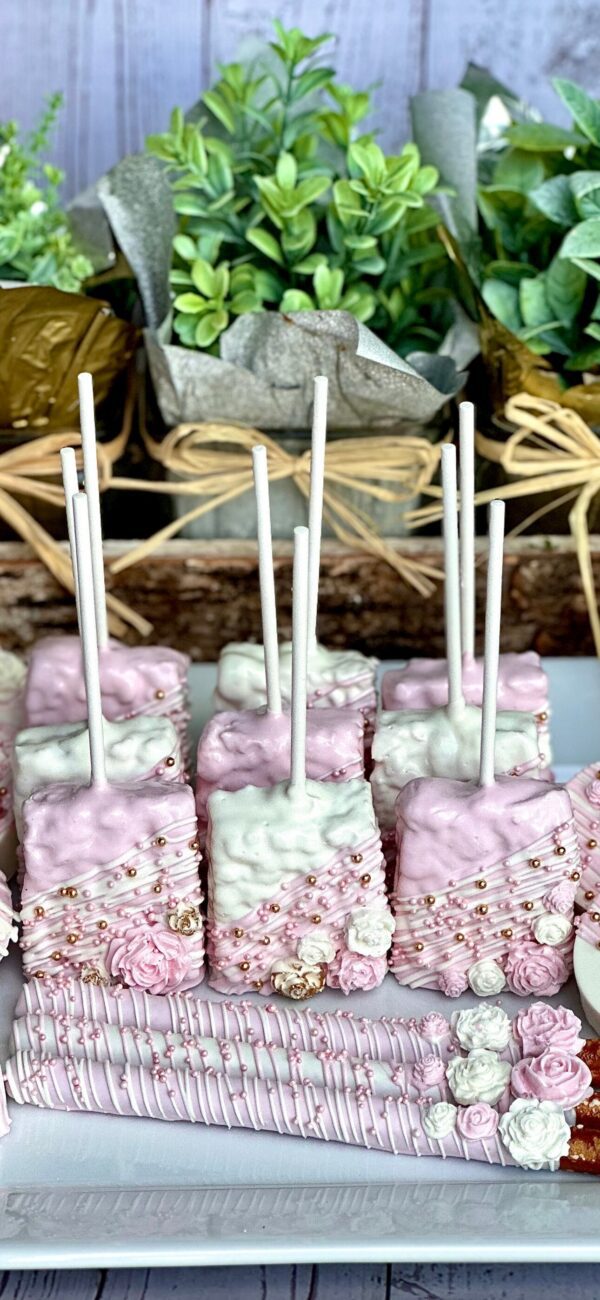 The Pink and White Chocolate Treat Party Bundle features marshmallows on sticks with pink and white decorations, pearls, and rose designs, elegantly arranged on a tray against a backdrop of greenery. Perfect for wedding, birthday, or baby shower favors.
