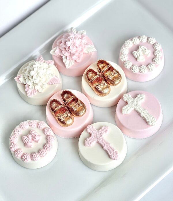 A white plate features seven decorated cookies in light pink and white, designed with flowers, ribbons, shoes, and crosses from the 12-Pink & White Chocolate Covered Oreos for Baptism/Christening/Holy Communion Favors.