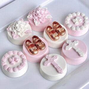 A set of eight decorated cookies with floral designs, pink bows, crosses, and gold baby shoes sits on a white tray, resembling the elegant style of the 12-Pink & White Chocolate Covered Oreos made for baptism or christening favors.