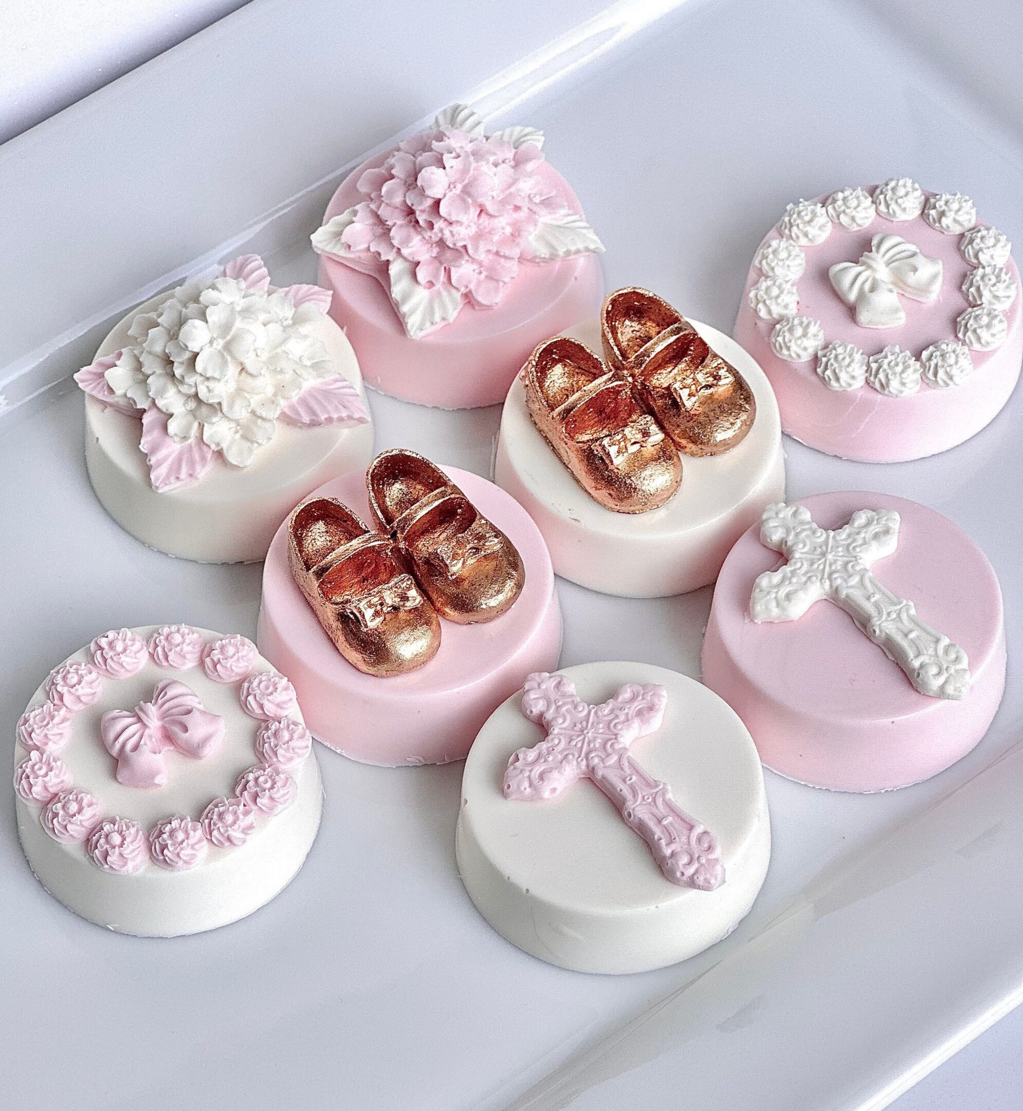 A set of eight decorated cookies with floral designs, pink bows, crosses, and gold baby shoes sits on a white tray, resembling the elegant style of the 12-Pink & White Chocolate Covered Oreos made for baptism or christening favors.