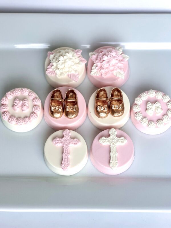 A white plate displays round cookies with pink and white icing, floral designs, gold baby shoes, bows, and crosses. These are "12-Pink & White Chocolate Covered Oreos For Baptism/Christening/Holy Communion Favors.