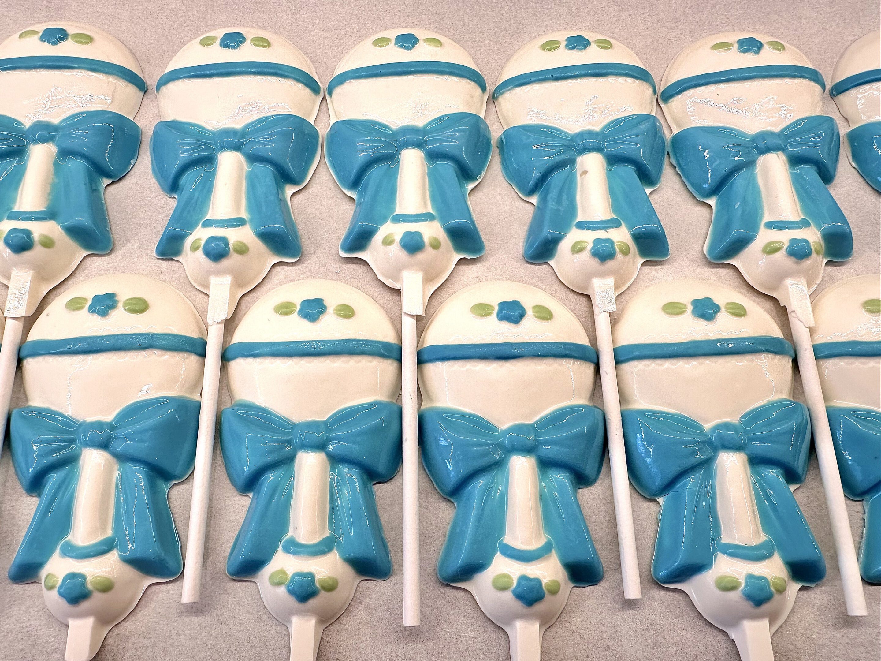 Rows of "12-Sweet Baby Boy White Chocolate Baby Rattle Lollipop Favors" featuring blue bows and decorative accents.