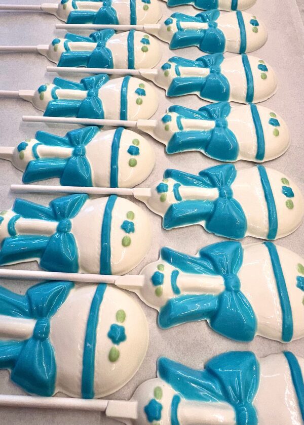 12-Sweet Baby Boy White Chocolate Baby Rattle Lollipop Favors, adorned with blue ribbons and green accents, are arranged in a grid for baby shower favors.