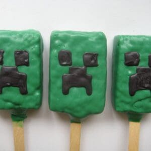Three pixelated treats on sticks, styled like Minecraft Creeper faces in green and black, are set against a white background; these are part of the 12-MineCraft Chocolate Covered Rice Krispie Treats.