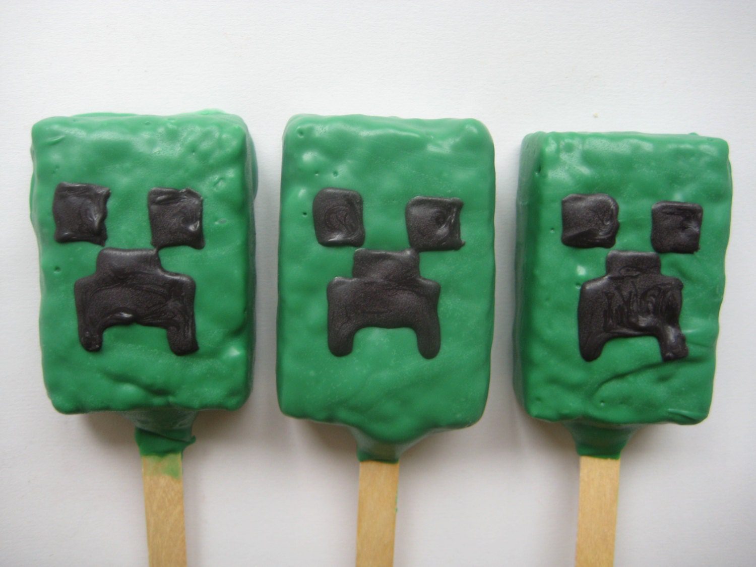 Three pixelated treats on sticks, styled like Minecraft Creeper faces in green and black, are set against a white background; these are part of the 12-MineCraft Chocolate Covered Rice Krispie Treats.