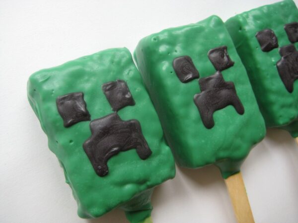 Three MineCraft chocolate-covered rice krispies treats on sticks, designed to look like pixelated green faces with black square eyes and mouths.