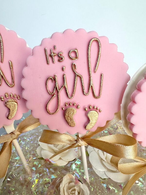 Pink lollipop with gold "It's a girl" lettering, gold baby footprints, and a gold ribbon. Product: 12 It's A Girl White Chocolate Lollipops for Baby Shower/Party Favors/Baby Sprinkle/Baby Favors.