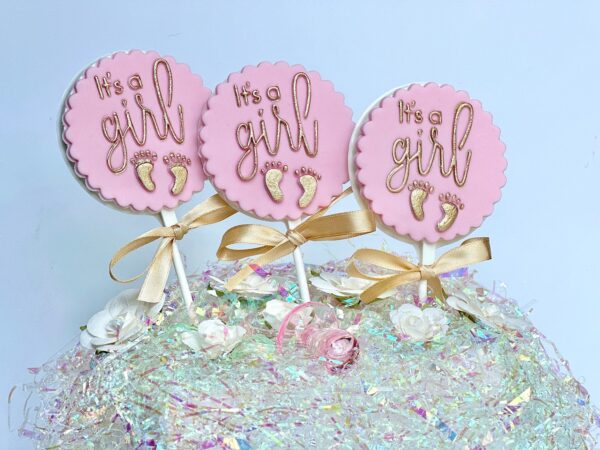 Three white chocolate lollipops labeled "It’s A Girl" sit on a decorative surface adorned with iridescent tinsel and white flowers.