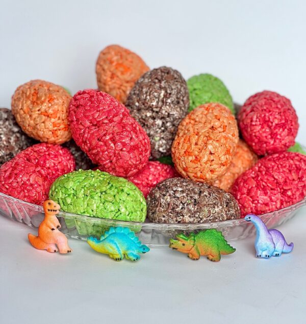 Displayed is a glass plate featuring the "12-Dinosaur Egg Rice Krispie Treats" for parties, accompanied by small dinosaur figurines in orange, blue, green, and purple.
