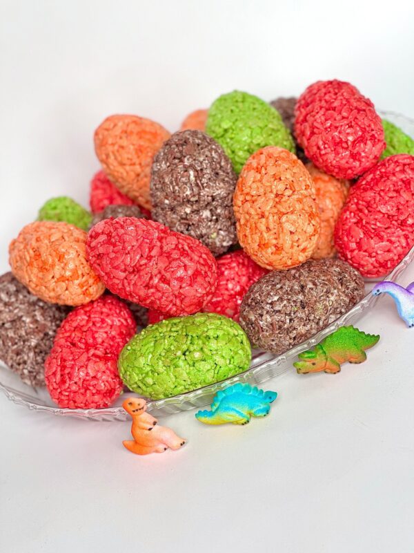 A glass plate features 12 colorful Dinosaur Egg Rice Krispie Treats, perfect for birthday parties, surrounded by small dinosaur figurines.