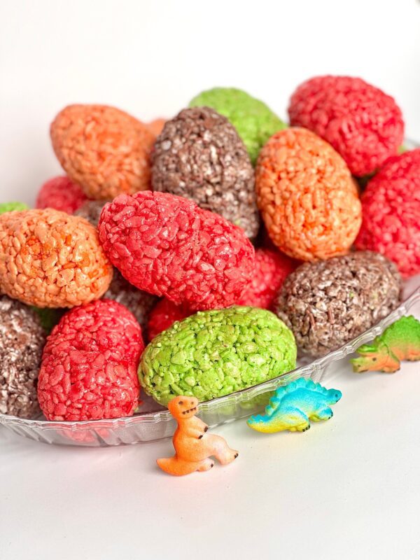 A plate of 12 Dinosaur Egg Rice Krispie Treats, perfect for birthday parties or dinosaur-themed events, adorned with small dinosaur figurines around the edge.