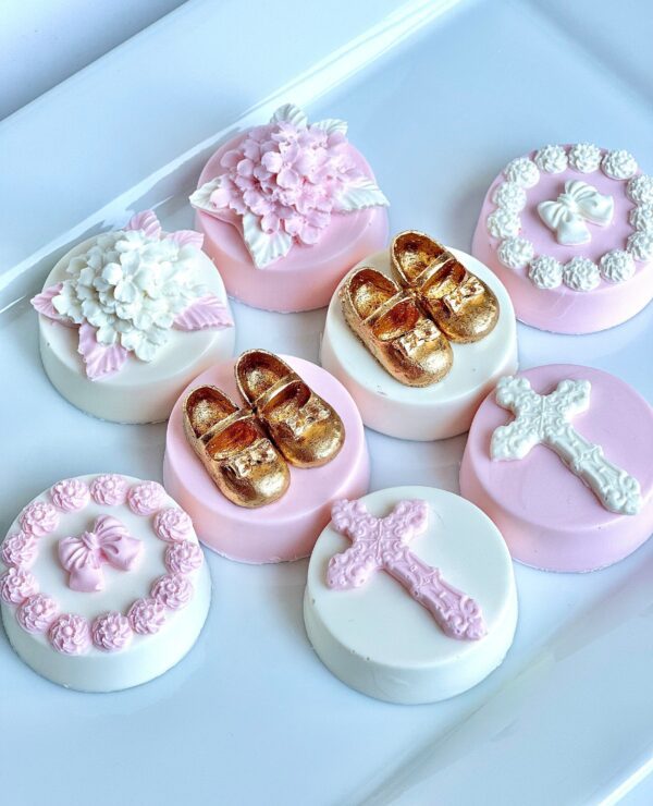 An assortment of 12 pink and white chocolate-covered Oreos, designed for baptism or Holy Communion favors, with gold shoes, crosses, floral designs, and bows on a white platter.