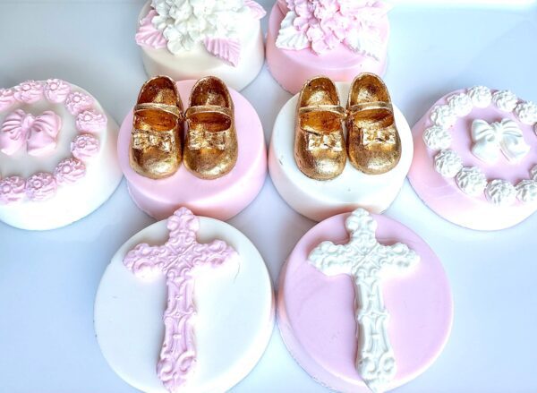 Decorative pink and white chocolate-covered Oreos for baptism, featuring gold shoes, floral designs, and white crosses.