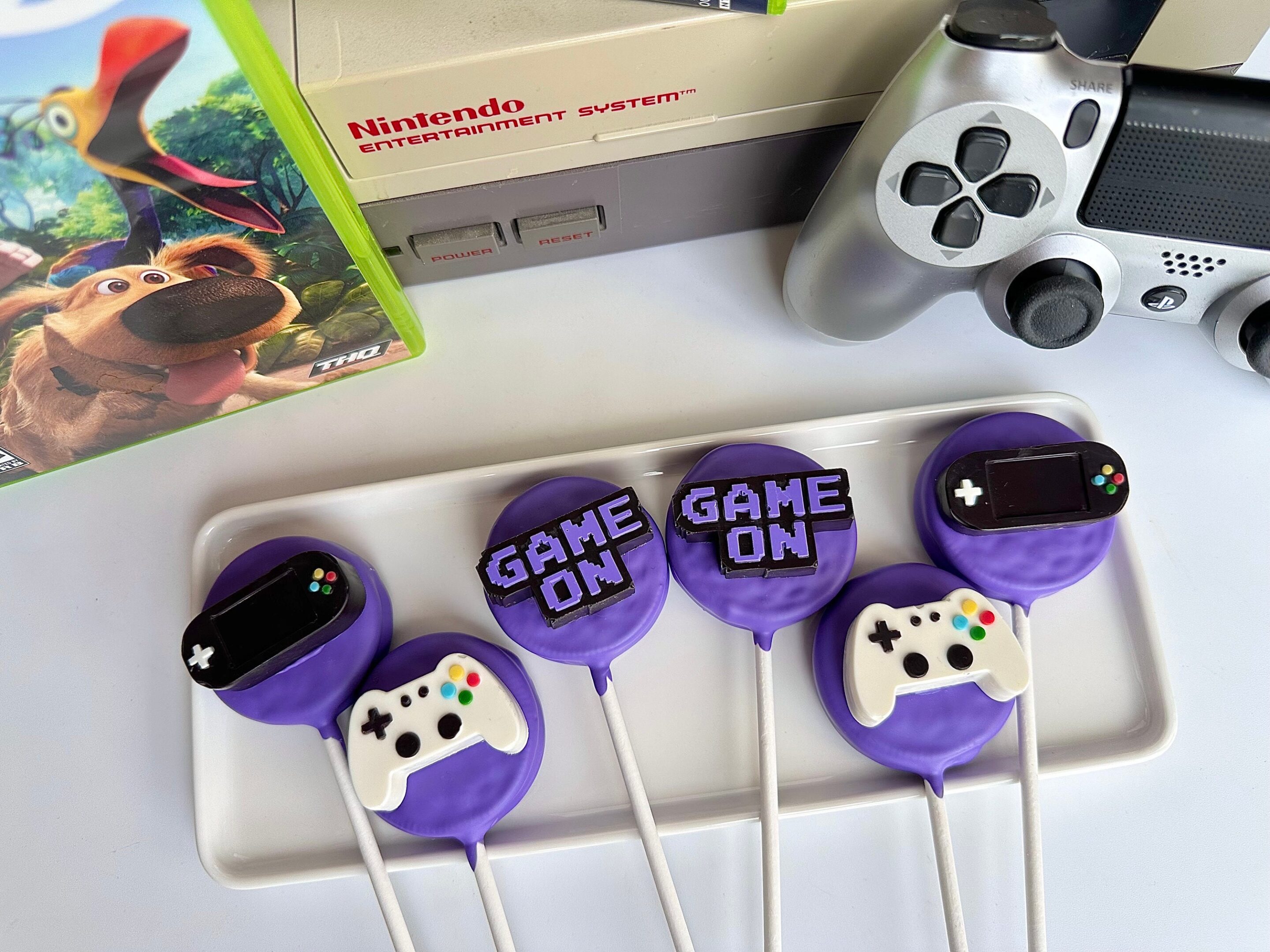 Displayed on a plate are purple "12-Video Game Controller Chocolate Covered Oreo Pops" with game controller designs and "Game On" text, while a Nintendo console, game case, and controller can be seen in the background.